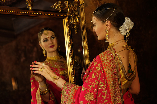 Female model Hindu Bride in saree, wearing gold and jasmine flower garlands in the hair looks in the mirror and touches his reflection, back view.