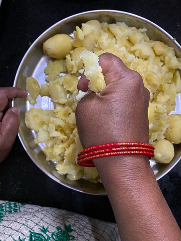 Stock photo showing the hand of an Indian woman making dinner by squeezing cooked boiled potatoes by hand, making masala mashed potatoes recipe of Hari Ghotra by using hands to squeeze and mash each potato on a silver thali plate, wearing red bangles.