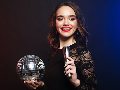 Lifestyle, party and people concept: young woman wearing black dress, holding disco ball and singing into microphone over dark background