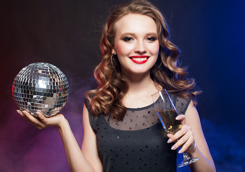 Holiday, people and celebrate concept: Beautiful woman in evening dresses, chic hairstyles and makeup holding a disco ball and glasses with champagne.