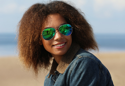 Beautiful young african american woman in a denim jacket and sunglasses on the beach on a summer day.