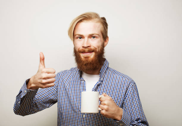 istock young man holding white cup with coffee or tea and show thumb up over grey background. 1405942103