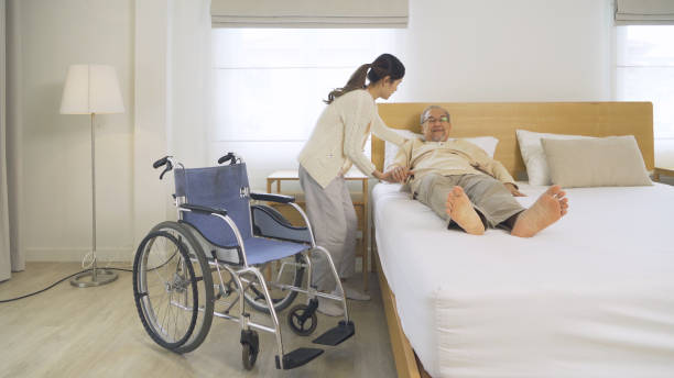 Asian woman nurse, daughter help father to get up from bed to wheelchair, supporting old senior elderly patient in bedroom in home or house in healthcare. People lifestyle. Family disability therapy. stock photo