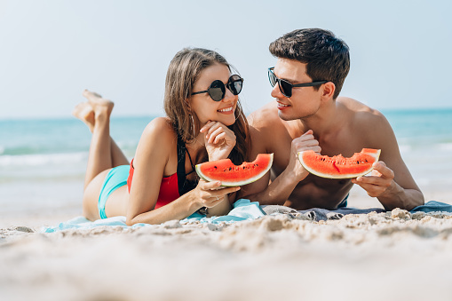 Happy young couple holding and eating slices of watermelon on the beach.Casual lifestyle, love, dating, Vacation concept.