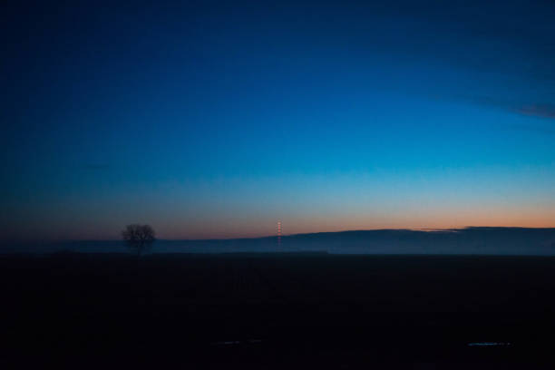 Winter sunrise, dark blue sky with orange sun coming up, silhouetted trees and mist