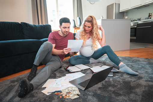 Caucasian male and pregnant female sitting on the floor and checking their finances together. They are using a laptop and reading documents.