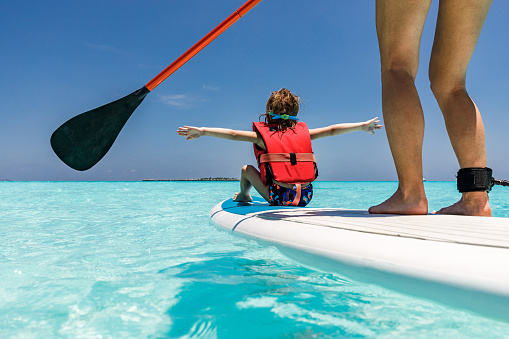 Rear view of carefree boy having fun with his arms outstretched on paddle board with his unrecognizable mother in summer day at sea. Copy space.