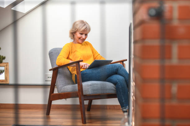 Senior Woman Reading News on Digital Tablet while Seated Usual afternoon routine. Senior woman reading news on digital tablet. She seats at home on a quiet corner. woman sitting cross legged in chair stock pictures, royalty-free photos & images