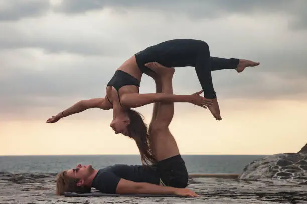 Fit young couple doing acro yoga for healthy lifestyle on tropical coast. Active woman balancing on partner feet, stretching at acroyoga pose. Sports exercises activity to restore strength and spirit