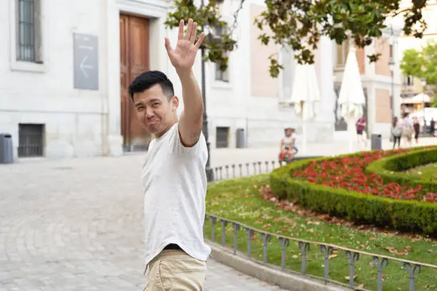 Asian man looking sad while waving hand to say goodbye to someone outdoors in the street.