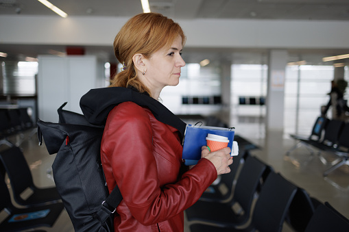 Female passenger waiting in airport departure area, holding traveling documents in hand and coffee to go