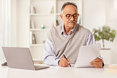 istock Mature man sitting in front of a laptop computer looking at a paper document 1405960956