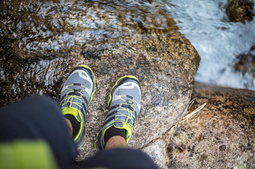 First person view of male legs in trekking sneakers at the edge of a stream in the mountains