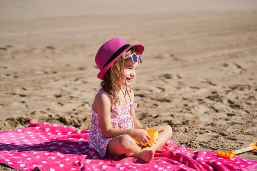 little girl sitting on towel with pink hat and sunglasses on the beach