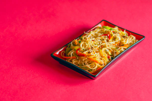 Traditional Korean starch noodles chapche with vegetables and beef in a red rectangular plate on a red background, side view, copy space.