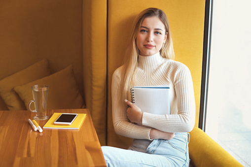 Portrait of beautiful young smiling  student girl sits in a cafe by the window and holding a notebook, looking at camera.