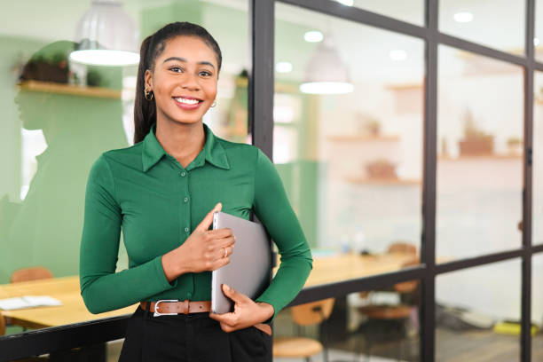 Carefree inspired african-american female employee standing in modern office space and holding laptop stock photo