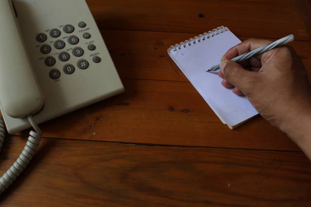 Hand is holding pen and writing in the notebook. telephone on the desk.