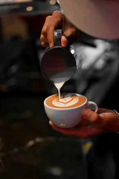 Photo of Professional male barista pouring a steamed milk into a coffee cup, making a latte art