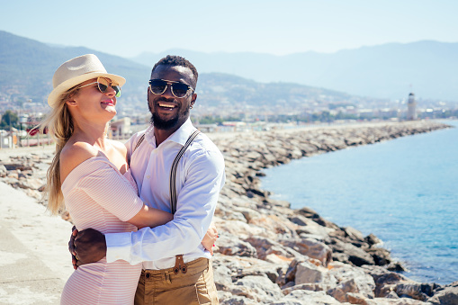 smiling happy european woman in straw hat and sexy pink dress walking with afro american ethnic man by summer beach with rock view in Turkey tropical resort.