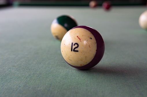 An old billiard ball with the number 12 (twelve) in lilac lies on the faded cloth of the billiard table.