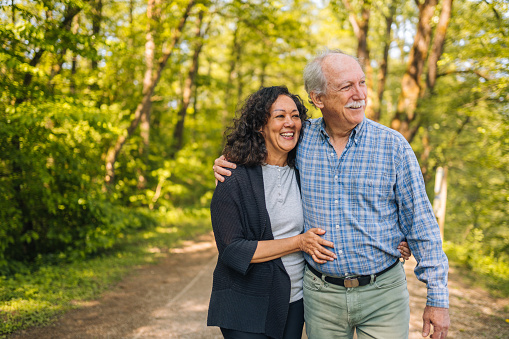Senior Hispanic lady taking a walk through a forest with a grey-haired, retired man, holding each other and smiling. Waist up image, natural sunlight and strong shadows.