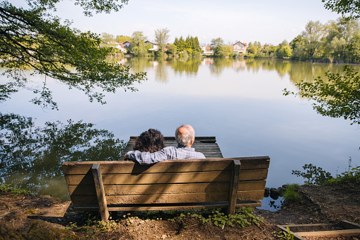 Back view of a grey-haired man holding a curly woman over the shoulders while sitting on a wooden bench next to a calm pond. Copy space. Only heads and shoulders visible.