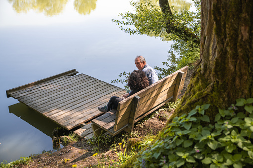 Side view of a grey-haired man and a a curly woman, sitting on a wooden bench next to a lake, a wooden platform in front of them. Copy space. Only man's face visible.