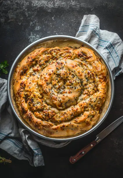 Turkish cuisine. Savory pie with Phyllo dough in baking pan. Su boregi, burek or borek on dark rustic background. Top view.