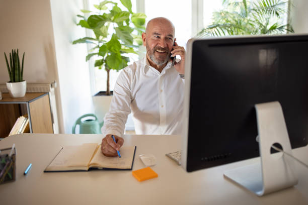 istock Mid-adult Caucasian man talking on mobile phone while working on computer from his modern home office 1405999006