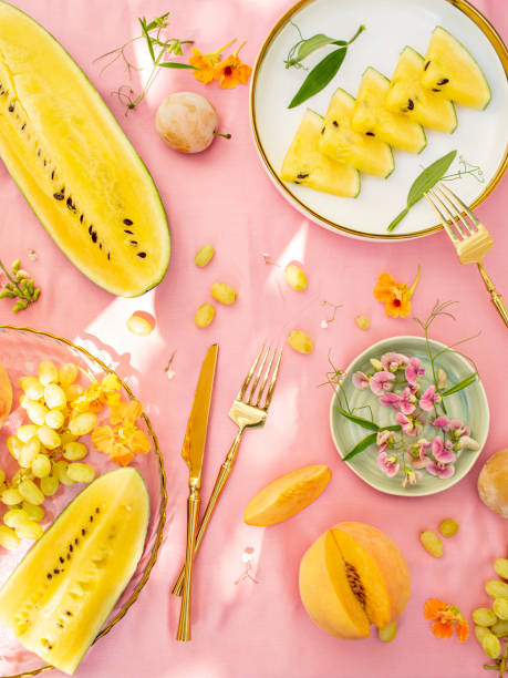 Sunny and bright open air breakfast with yellow watermelon, grape, plums and summer wild flowers decoration. Romantic picnic for couple. Floral botanical background, pattern.