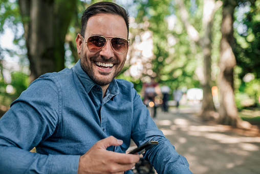 Portrait of happy businessman wearing sunglasses with smart phone sitting at park