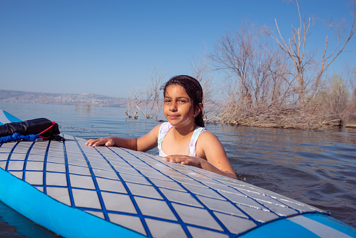 Portrait of an elementary-age girl resting before climbing on a windsurf board after falling in water. Windsurfer beginner kid windsurfing, sports training.  Active child and water sport. Windsurfing on summer vacation time on a sunny day on the lake.