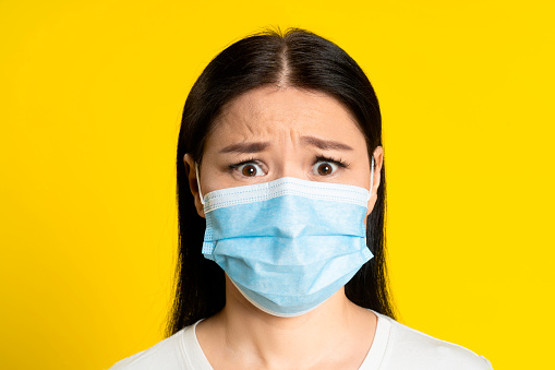 Scared of pandemic mature asian woman wearing medical face mask coronavirus or monkeypox prevention. Charming middle age woman in white t-shirt and medical mask on yellow background.