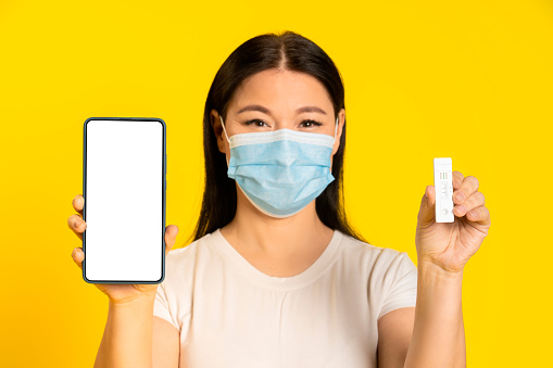 Happy asian woman wearing medical face mask, holding smartphone and test, screen showing app interface, health, vaccination passport, certificate. Call your doctor concept. Product placement mock up.
