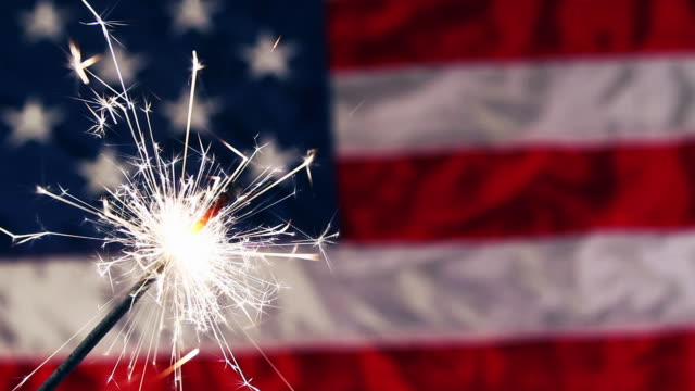 a sparkler firework with a USA Flag in the background.