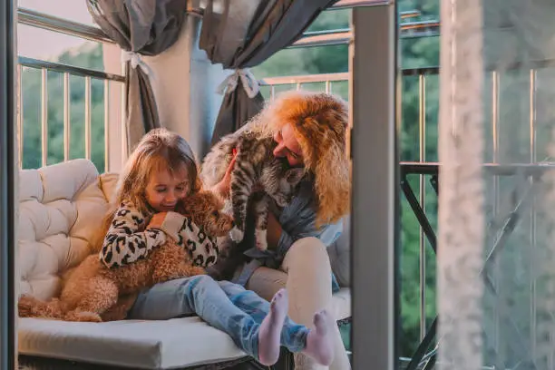 Photo of a little, happy girl, a child with a smile plays on a summer, spring vernada at home with mom and a dog at sunset. family weekend evening. Cat, dog, pets as a family member.
