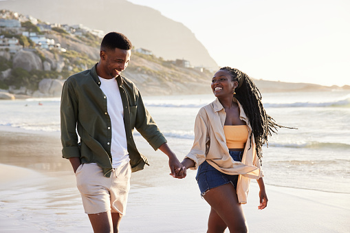 Laughing young African couple walking hand in hand together along a sandy beach on a late afternoon in summer