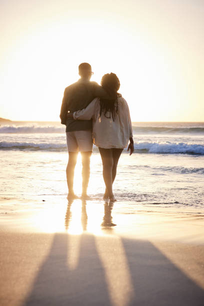 Loving young couple standing on a beach and watching the sunset Rear view of an affectionate young African couple standing arm in arm together on a beach and watching the sunset over the ocean black couple sunset stock pictures, royalty-free photos & images
