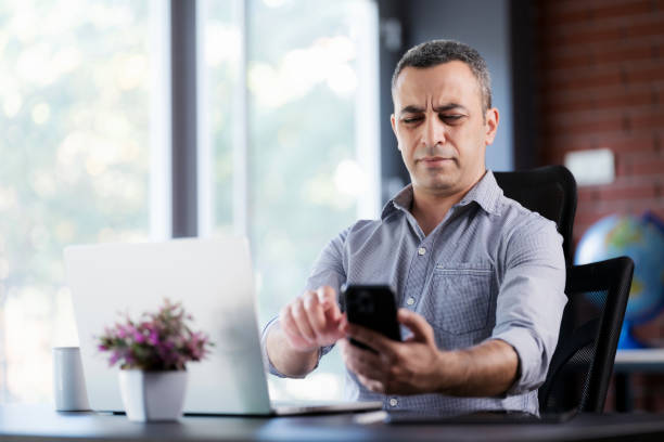 Tired Businessman In The Office Has Difficulty To See Texts On His Smartphone, Concept For The Beginning Of Presbyopia Just After The Age Of 40 Years. stock photo