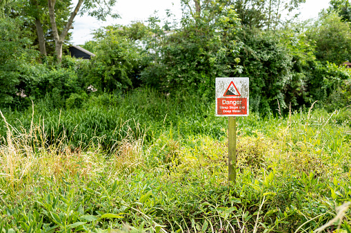 Step slow and deep water sign seen at the edge of a dangerous river bank, seen heavily overgrown which causes further risks.