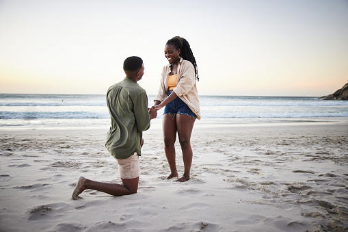 Young African man kneeling in the sand and proposing to his smiling girlfiend on a beach at dusk