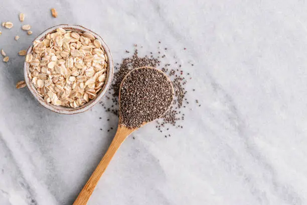 Overhead view of chia seeds on a wooden spoon and uncooked oat flakes in a small ceramic bowl on a grey marble background with copy space on the right side