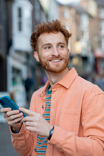 A young man spending the day in Durham, England and exploring the city. He is standing on a city street, looking to his side and smiling while using his mobile phone.