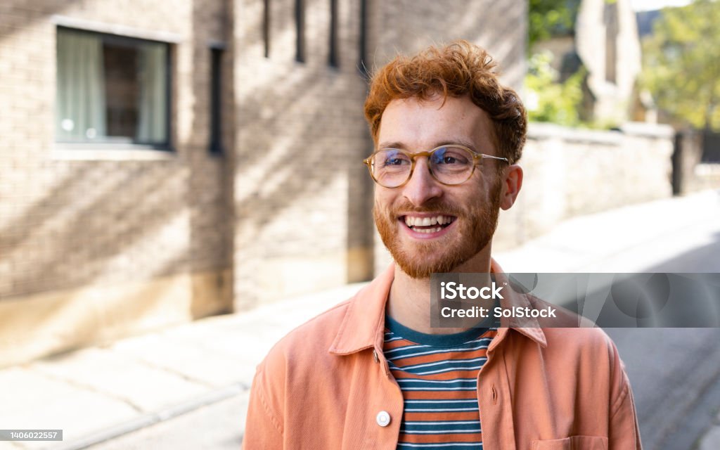 A Confident and Happy Young Man A young man spending the day in Durham, England and exploring the city. He is standing on a city street while looking to his side and smiling. Redhead Stock Photo