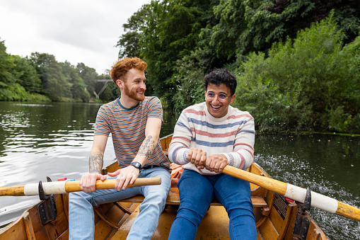 A male couple spending the day in Durham, England together. They are sitting in a rowboat and using one oar each to row the boat along the river. One man is looking at his fiancé and smiling while they row and the water is splashing around them.