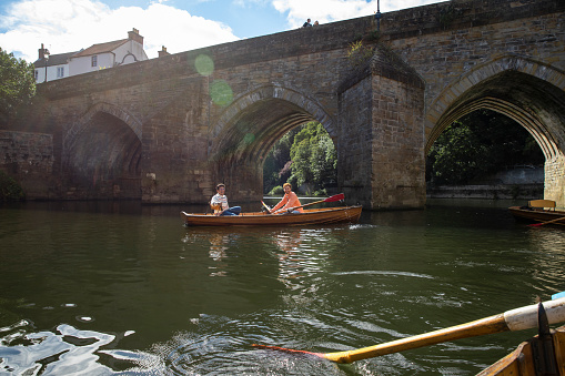 A male couple spending the day in Durham, England together. They are sitting in a rowboat and rowing along the river in the sun while looking at the camera and smiling.