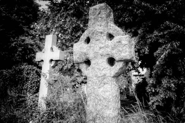 Ancient Celtic cross gravestone showing its ancient, heavily weathered texture seen with other graves in a European cemetery.