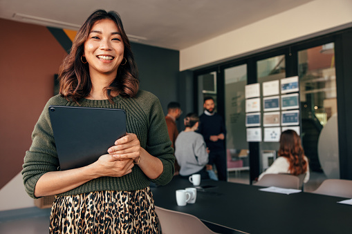 Cheerful young businesswoman smiling at the camera while holding a digital tablet. Happy young businesswoman standing in a boardroom with her colleagues in the background.