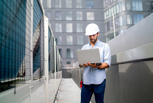 Caucasian engineer or technician worker use laptop to work in way on terrace of construction site with high building as background.